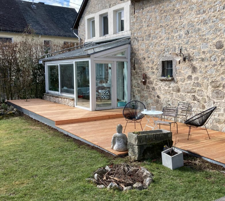 Gartenansicht eines Hauses aus Naturstein mit Wintergarten und Terrasse mit Holzbelag
