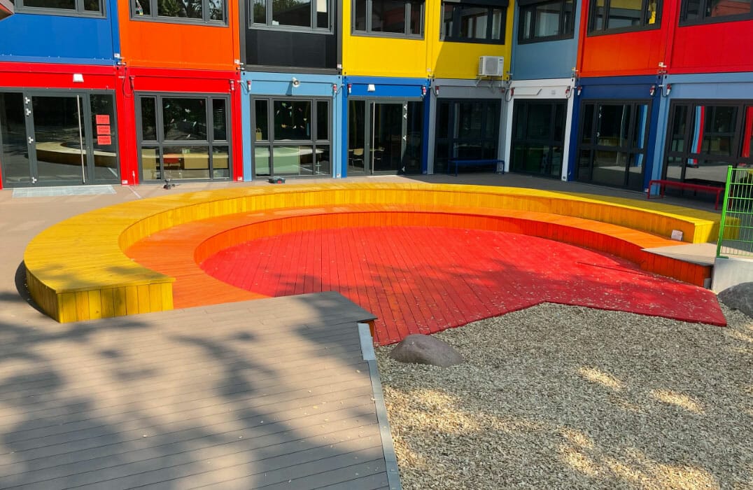 Ausschnitt eines farbenfrohen Schulgebäudes mit Sitzmöglichkeit und Platz aus Holz in rot, orange und gelb